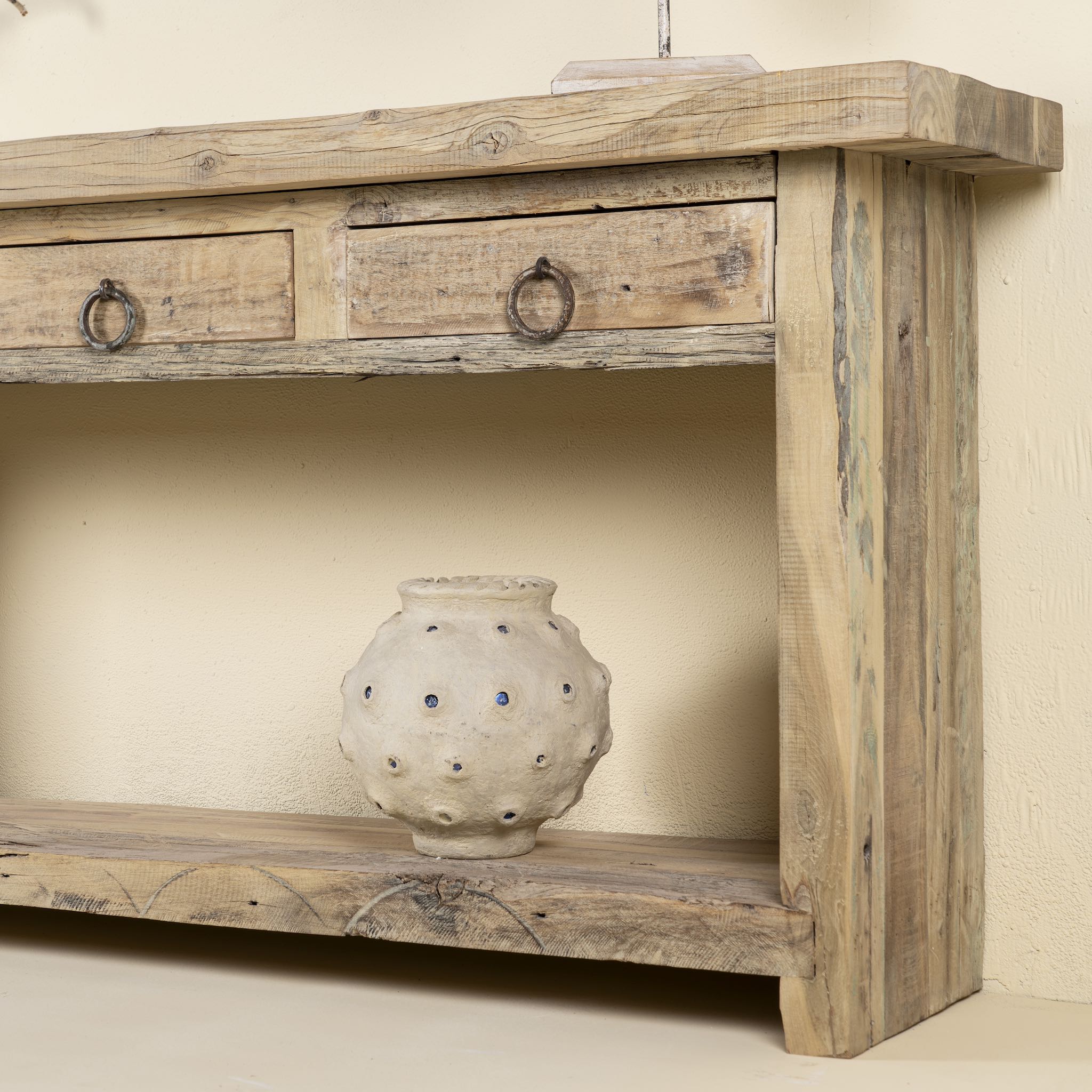 Close up side angle image of a rustic console table crafted from reclaimed teak wood, featuring a natural weathered finish, three drawers with metal ring pulls, and a lower shelf for additional storage or display, blending simplicity with organic charm.
