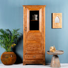 An antique Indian teak wood showcase with a glass door and bronze-colored hardware, featuring interior shelves and a drawer, placed against a blue wall next to a potted plant and a decorative mirror.
