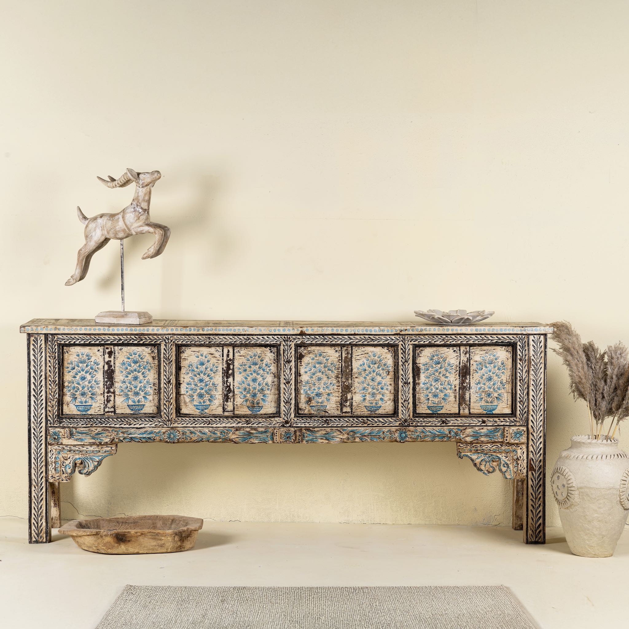 Front view image of a hand-painted console table made from reclaimed teak wood, featuring intricate floral motifs in blue tones across the front panels, framed by carved detailing and a distressed finish that highlights its artisanal and vintage character.

