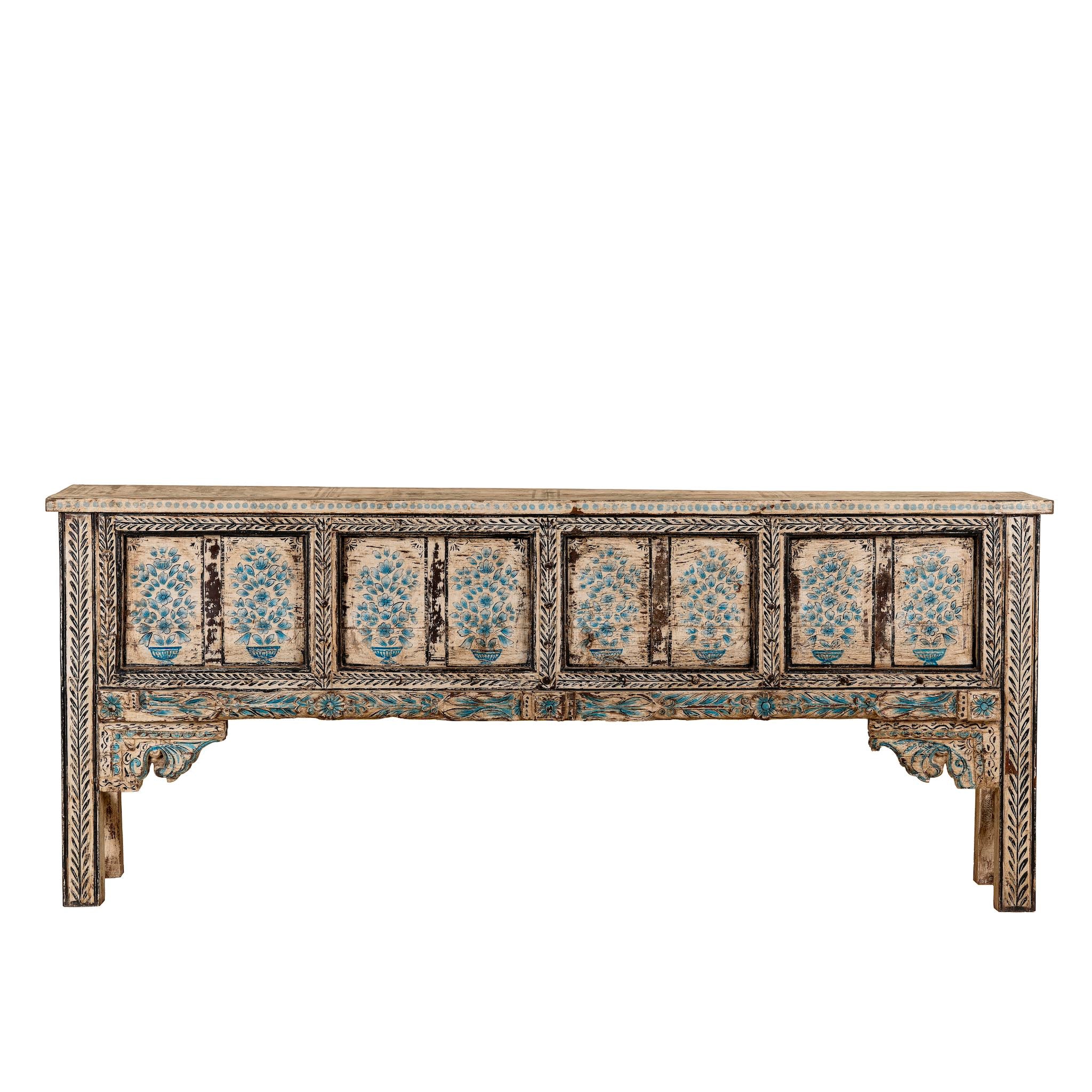 White background image of a hand-painted console table made from reclaimed teak wood, featuring intricate floral motifs in blue tones across the front panels, framed by carved detailing and a distressed finish that highlights its artisanal and vintage character.
