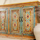 Side angle close up image of a hand-painted sideboard made from reclaimed wood, featuring four intricately detailed doors with vibrant floral and geometric motifs in soft blues, yellows, and reds, finished with a distressed vintage patina for a charming, artisanal look.
