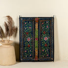 Hand-painted wall panel made from an old teak wood window frame, adorned with detailed floral motifs in shades of pink, red, green, and white against a deep blue background, framed by intricate carvings and colourful decorative accents.
