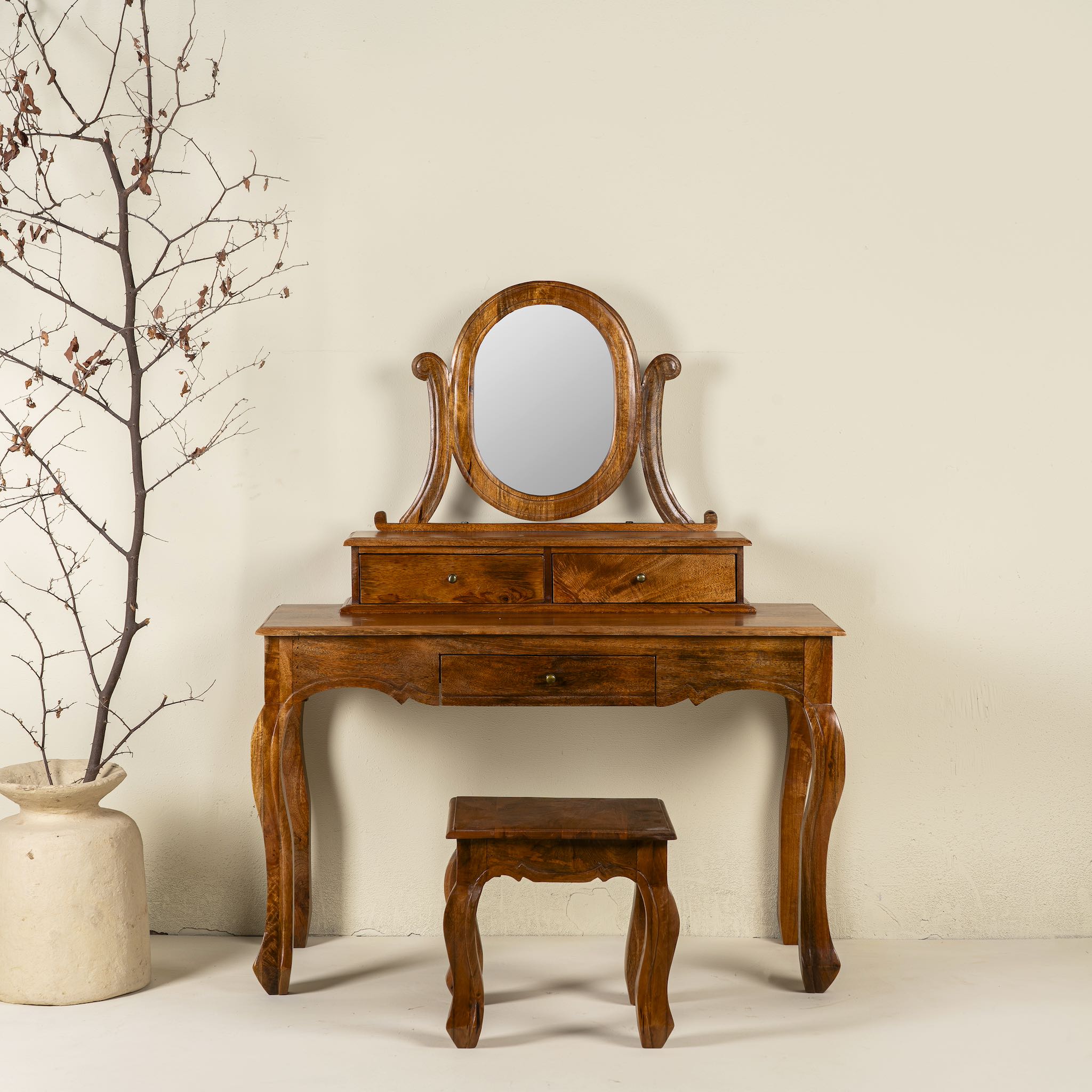 A handcrafted mango wood cosmetic table with a curved mirror and a matching chair, placed against a neutral background with a decorative branch in a vase to the side.