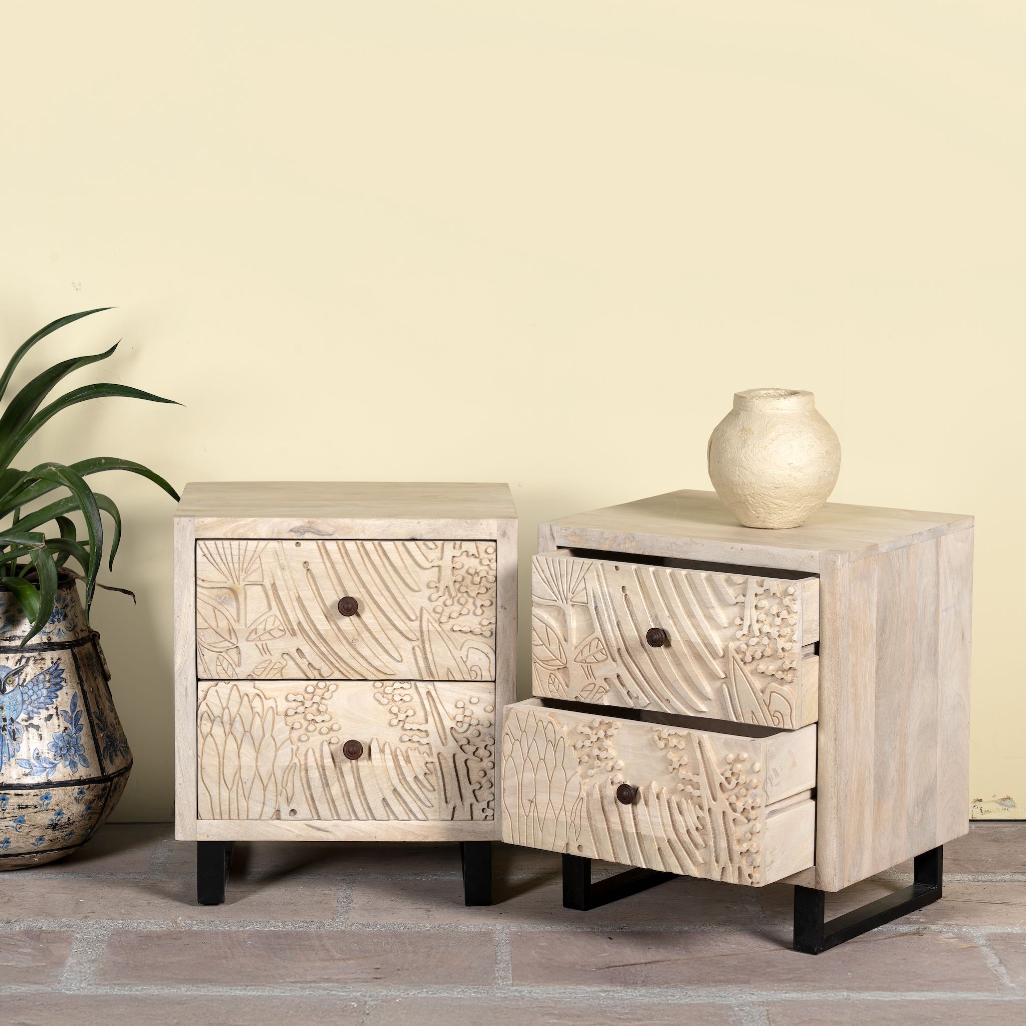 Side angle image with open drawer of a pair of mango wood nightstands with hand-carved sandblasted finish and black iron stand; artisanal bedroom furniture, 51 × 41 × 59 cm.