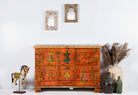 Hand painted teak wood storage chest with vibrant orange color and traditional Indian patterns, displayed on a white background with decorative items around it.