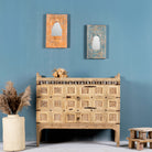 An antique Indian treasure chest with sandblasted surface and hand-carved patterns, featuring iron applications, displayed against a blue wall with decorative items and a vase beside it.