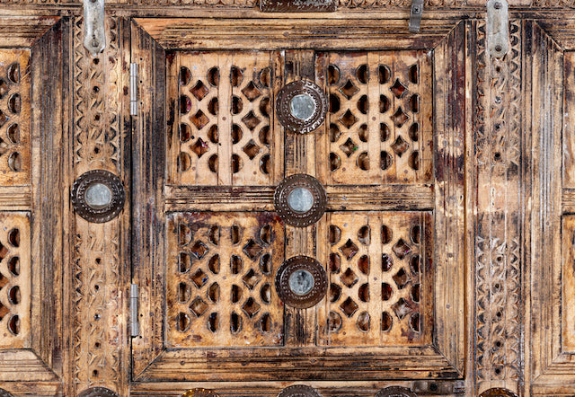Close up image of an antique Indian treasure chest in solid teak with sandblasted finish, hand-carved details, and iron accents. Vintage home accessory for eclectic Indian decor, 127 × 110 × 47 cm.