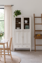 lifestyle image of a shabby chic teak wood cabinet with glass doors, drawers, and compartments – sustainable living room furniture crafted from refurbished teak wood with a soft white finish.