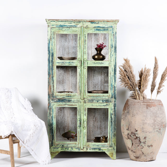 A refurbished teak wood showcase with a distressed vintage finish in green, featuring glass panelled doors and multiple shelves, placed next to a large vase with dried plants.