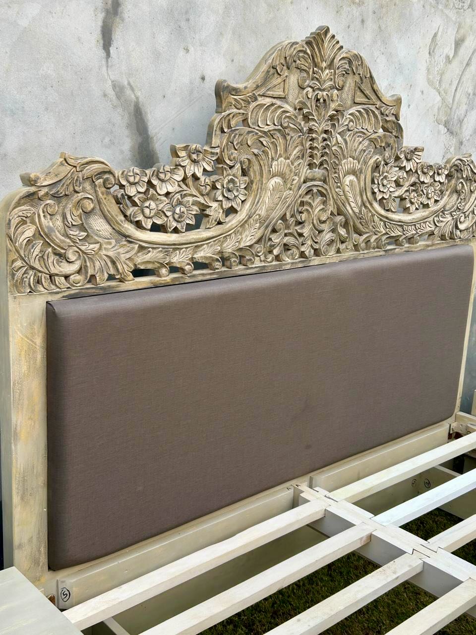 Close up of the head board of a hand-carved vintage bed Samar made from distressed mango wood, featuring an ornate high headboard with soft grey fabric upholstery. A unique blend of Indian craftsmanship and contemporary comfort in a natural, neutral palette.