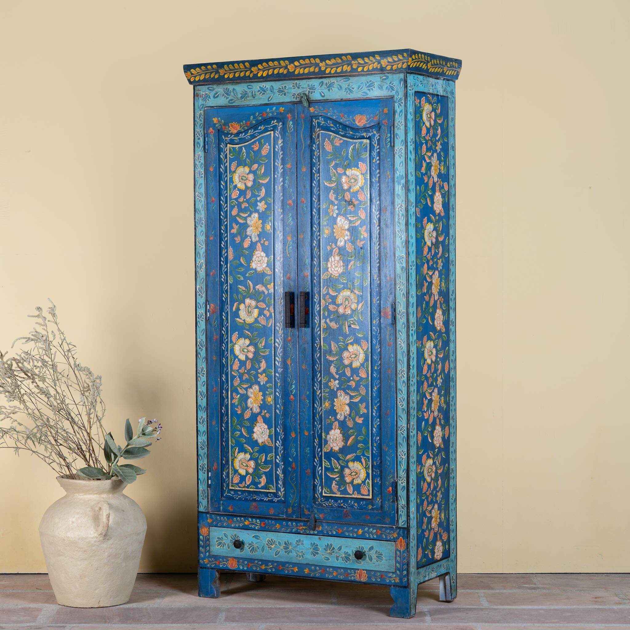 Side angle image with closed doors o a hand-painted wardrobe made of reclaimed wood in vibrant blue floral design – Yuvika model by Om Home & Garden