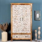 Hand-carved mango wood wardrobe with whitewashed floral carvings and a natural wood body, featuring three doors and a drawer, displayed in a room setting with decorative items around it.