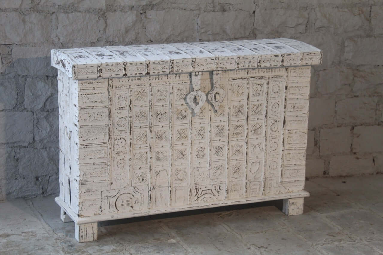 An antique white-colored box with brass fittings and hand-carved patterns, placed against a stone wall background.