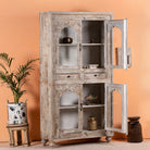 Side angle image with open door of a hand-carved mango wood showcase with glass doors and drawers – carved wooden furniture in distressed turquoise and blue, combining artisan detail with vintage charm.